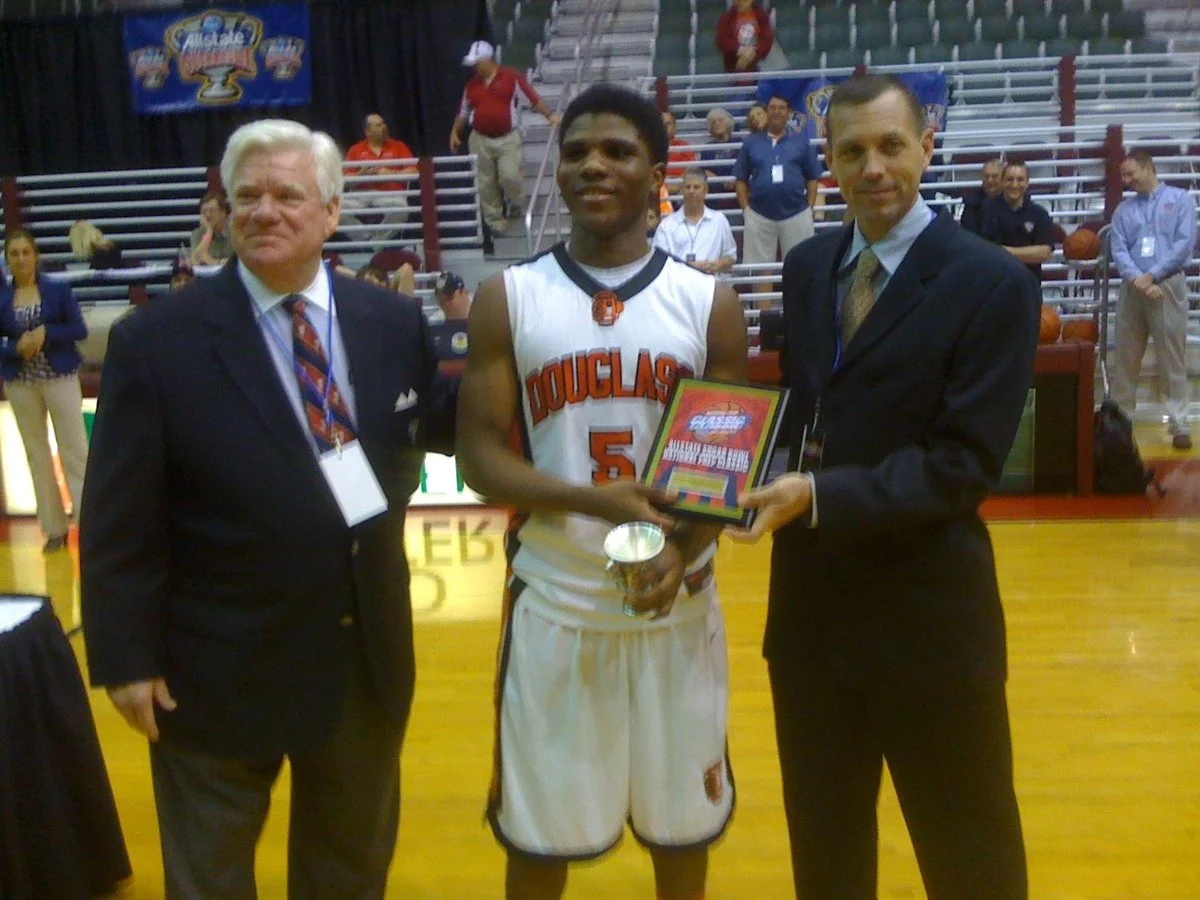 A young basketball player in a Douglass jersey holding a trophy and certificate, flanked by two men in suits, during an award ceremony in a gymnasium.