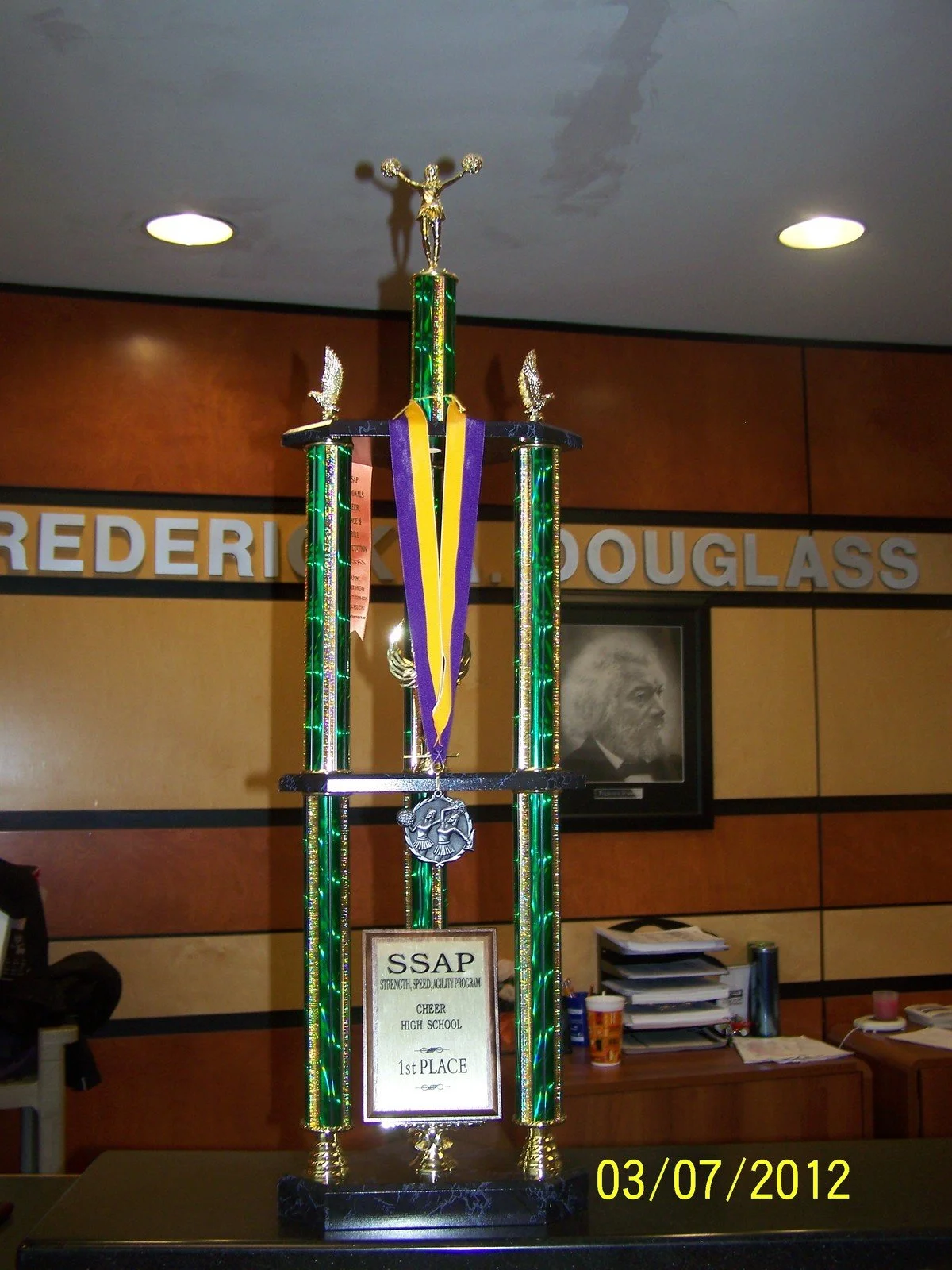 A large trophy with a gold figure of a cheerleader on top, four green columns, and various medals hanging from it, placed on a table in front of a mottled brown and beige wall with a plaque of a man's profile and a sign that reads 'FREDERIC K. DOUGLA