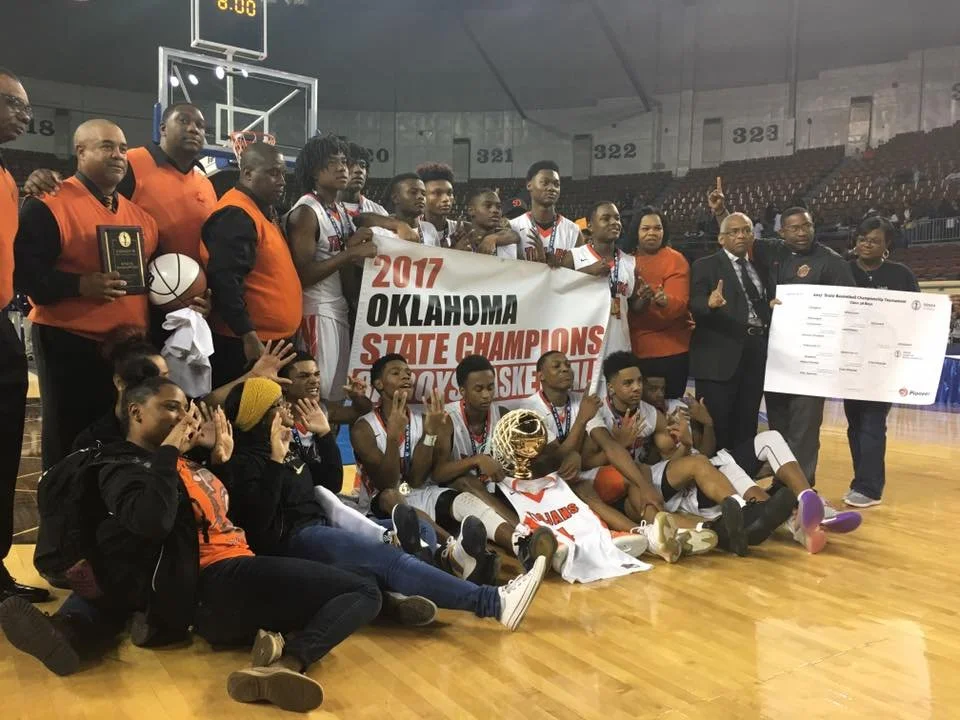 Oklahoma high school boys basketball team and supporters celebrating with a banner that reads '2017 Oklahoma State Champions Boys Basketball' inside a basketball arena, holding a trophy and a large check.