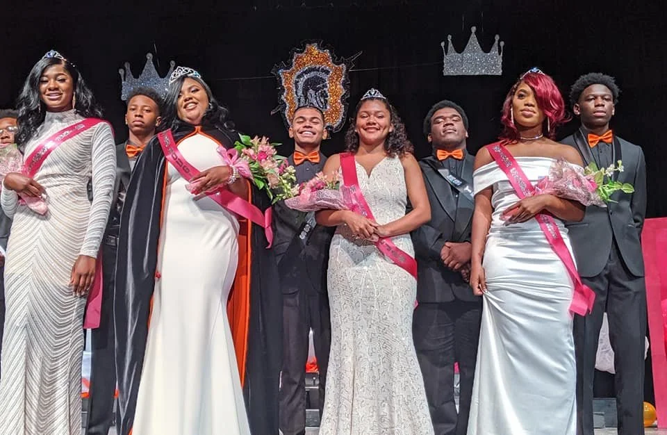 Group of young people at a pageant, wearing formal dresses and suits, with crowns and sashes, holding flowers, smiling on stage with a decorated backdrop.