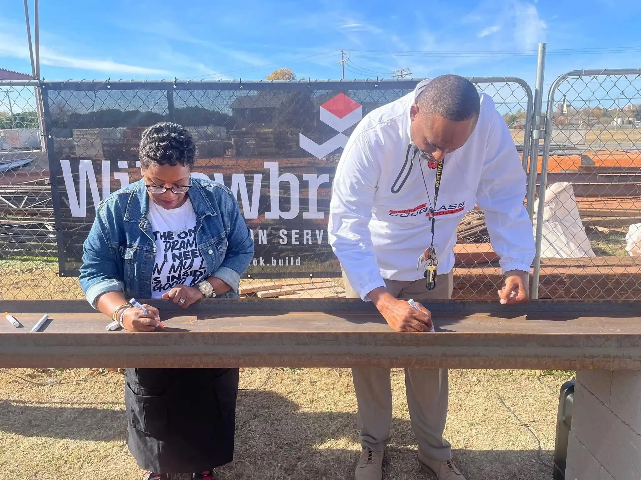 Two people signing a large wooden beam outdoors, with construction fencing and a sign behind them.