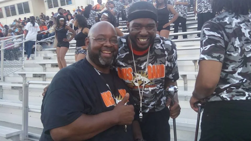 Two smiling men at a sports event, one with a beard and glasses, giving a thumbs-up, and the other with a cane and wearing a bandana, surrounded by people in athletic clothing