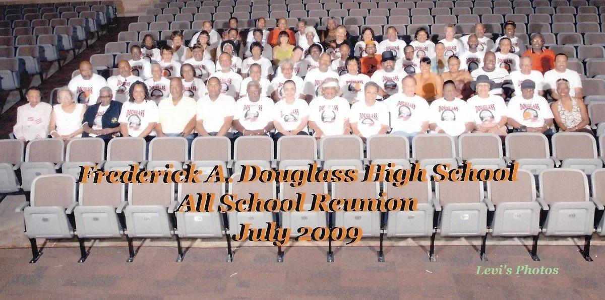 A large group of people sitting in an arena-style seating area, attending a reunion event for Frederick A. Douglass High School in July 2009. Many are wearing white T-shirts with a logo or image, indicating they are part of the class reunion. The tex