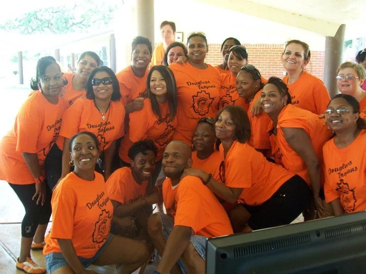 Group of women and one man wearing orange T-shirts, posing together indoors in front of a window, smiling for a group photo.