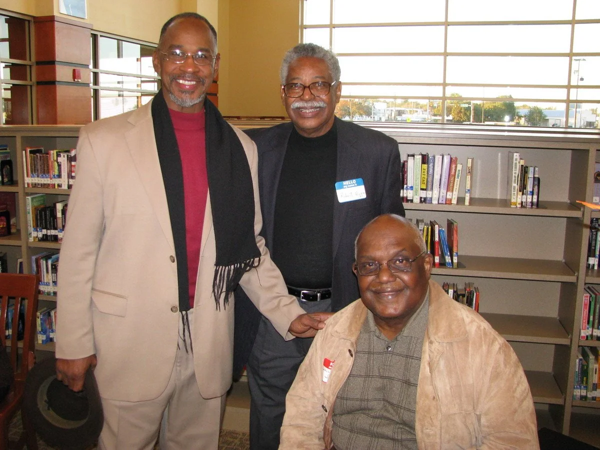 Three men in a library, two standing and one seated, smiling at the camera, with bookshelves in the background.