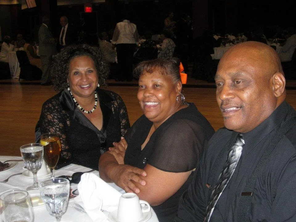Three people are sitting at a banquet table, smiling. They are dressed in formal attire, and there are drinks and tableware on the table. The setting appears to be a social event or celebration in a large hall.