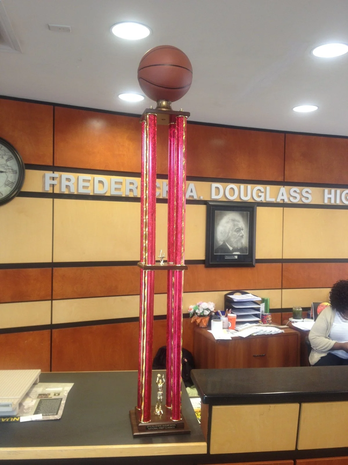Tall basketball trophy with a basketball on top, placed on a desk in front of a wall with a sign reading 'FREDERICK A. DOUGLASS HIGH' and a portrait of Frederick Douglass.