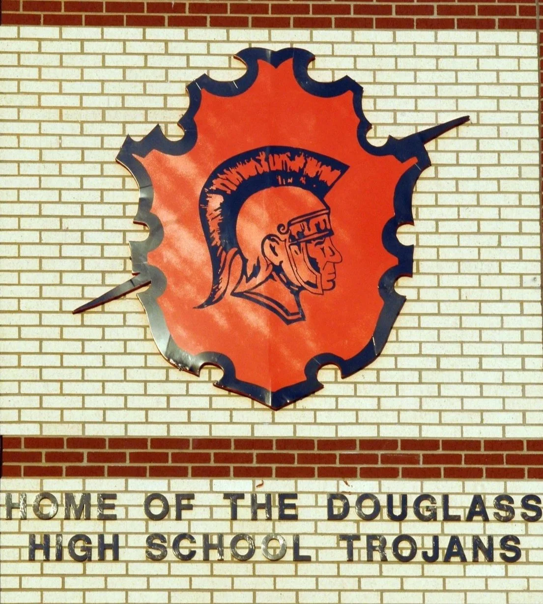 A school sign featuring a large, stylized Spartan helmet logo on an orange background, mounted on a brick wall, with the text "Home of the Douglass High School Trojans" below.