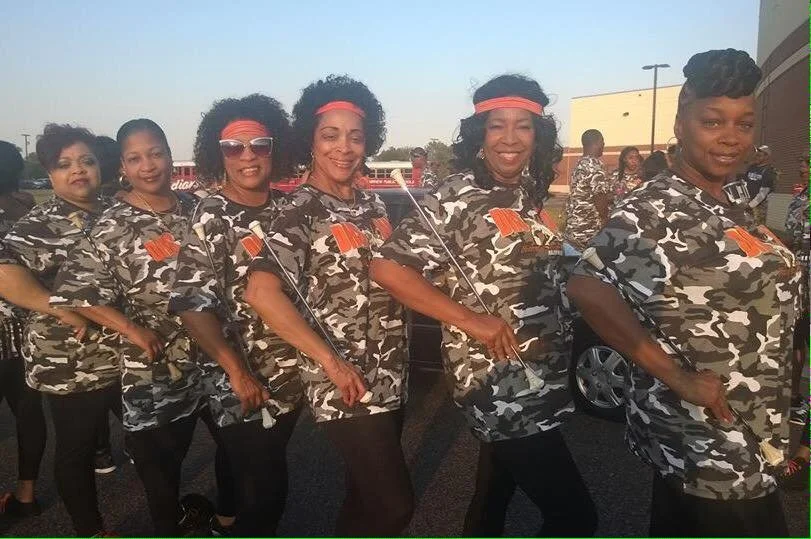 A group of women standing outdoors, dressed in matching camouflage outfits with orange ribbons and some wearing headbands and sunglasses, smiling at the camera.