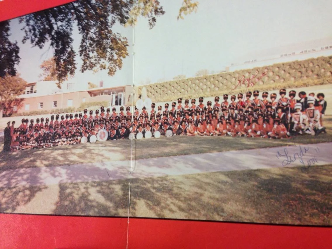 A large group of children dressed in band uniforms, posing for a photo outdoors on a grassy area with trees and a building in the background.