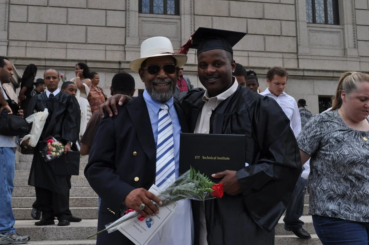 A man in graduation gown and cap standing next to an older man in a suit, both smiling; the graduate holds a diploma and a red rose. They are outside a stone building with steps, surrounded by other graduates and people.