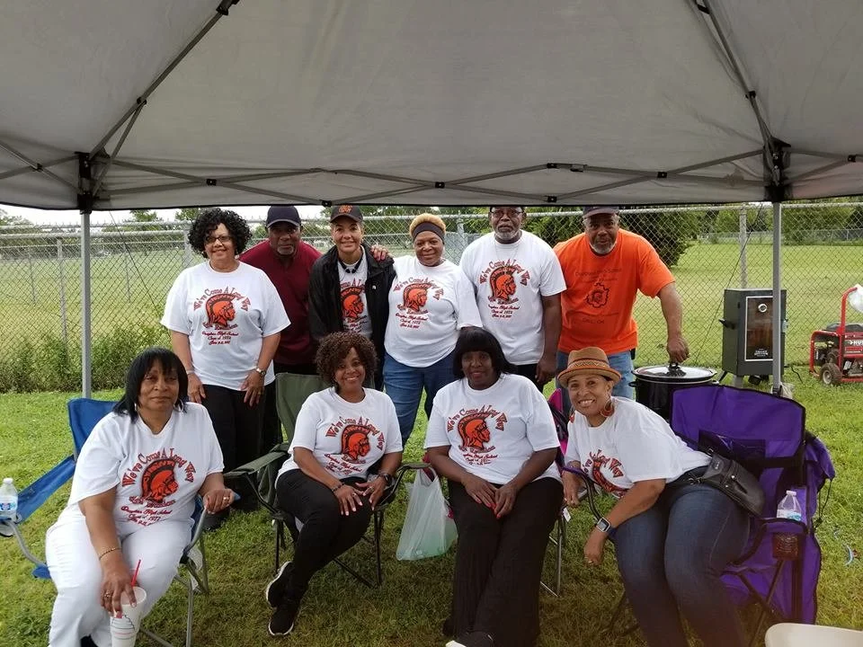 Group of nine people gathered outdoors under a canopy, some wearing matching white t-shirts with a red and black logo and text, on a grassy area with a chain-link fence and trees in the background.
