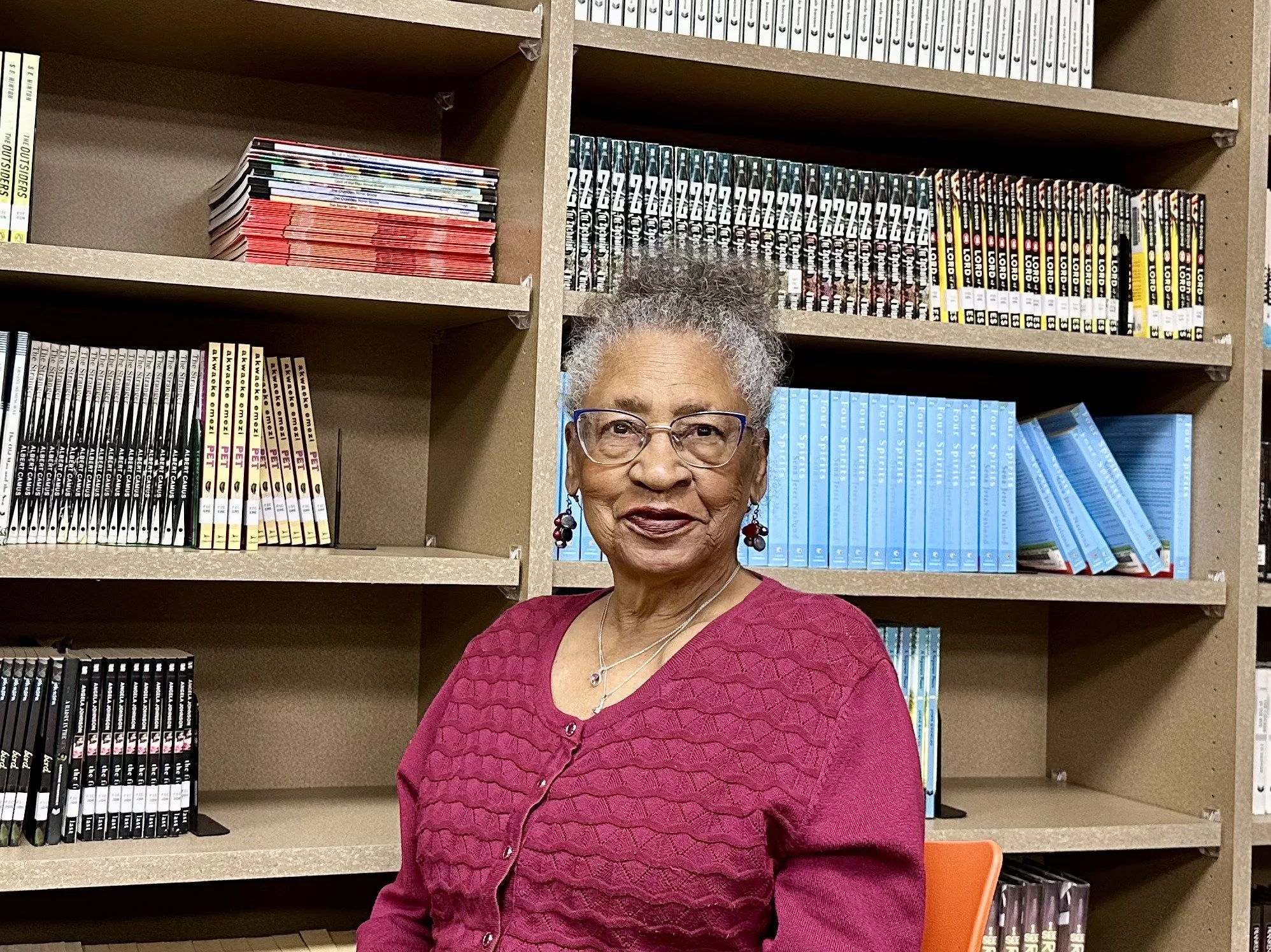 An elderly woman with gray hair, glasses, and earrings, wearing a pink sweater, sitting in front of a bookshelf filled with colorful books.