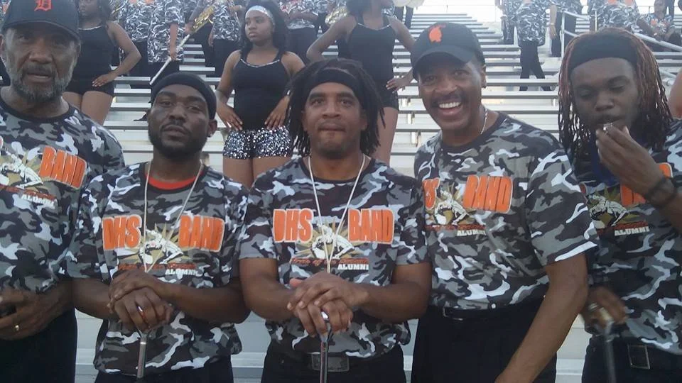 Group of five men in camouflage t-shirts with orange text and graphics standing together at a sports stadium, with women in similar attire in the background.