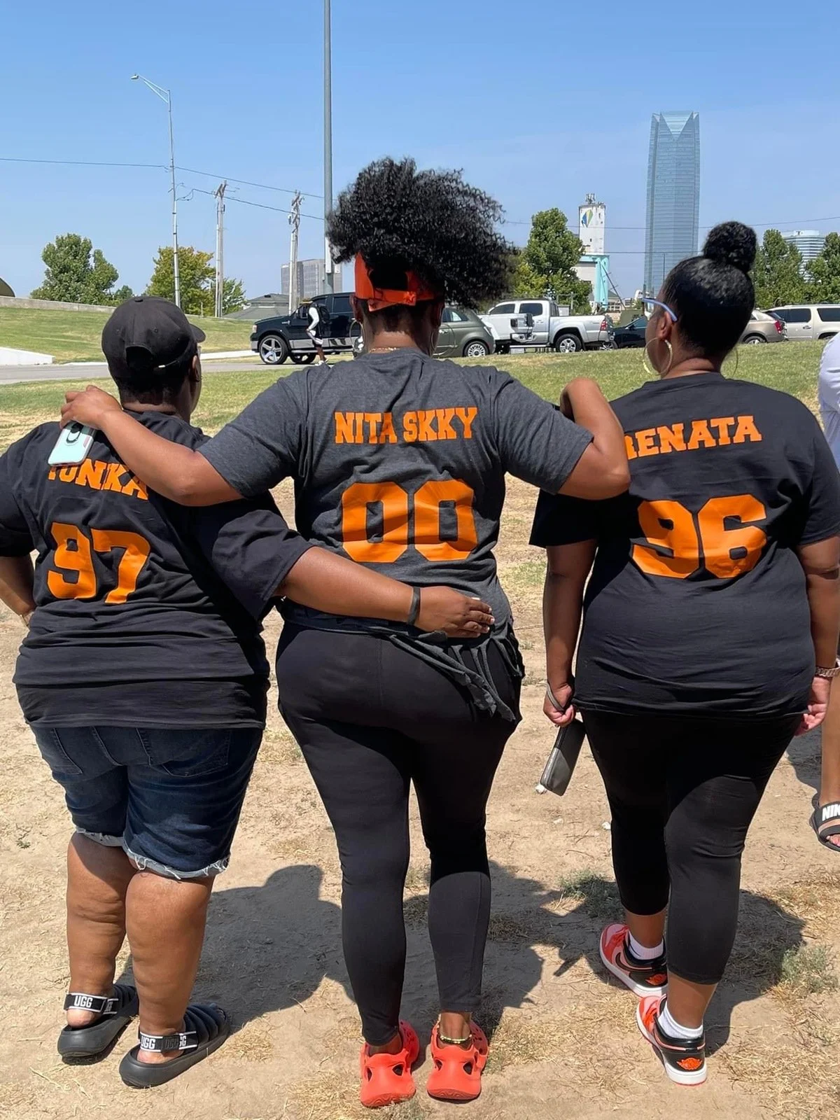 Three women with their arms around each other, wearing black T-shirts with orange letters and numbers, standing outdoors on a sunny day.
