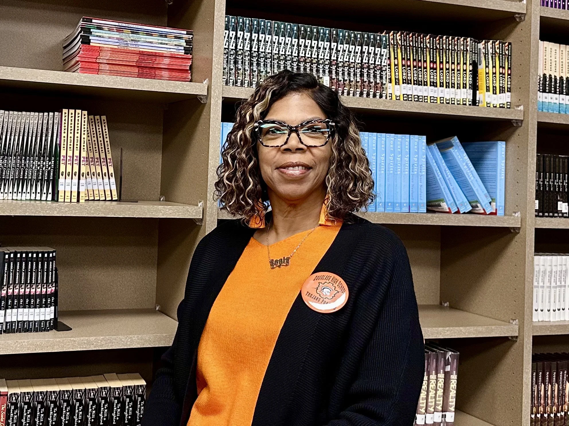 A woman with curly hair and glasses stands in front of bookshelves filled with books in a library.