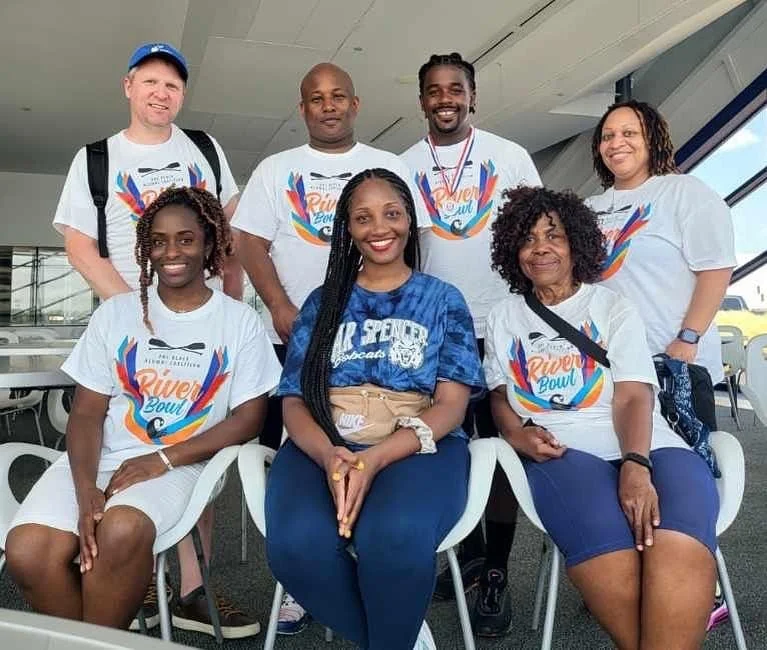 Group of seven diverse people posing for a photo at an event, some wearing 'River Bowl' t-shirts, indoors in a modern space with windows.