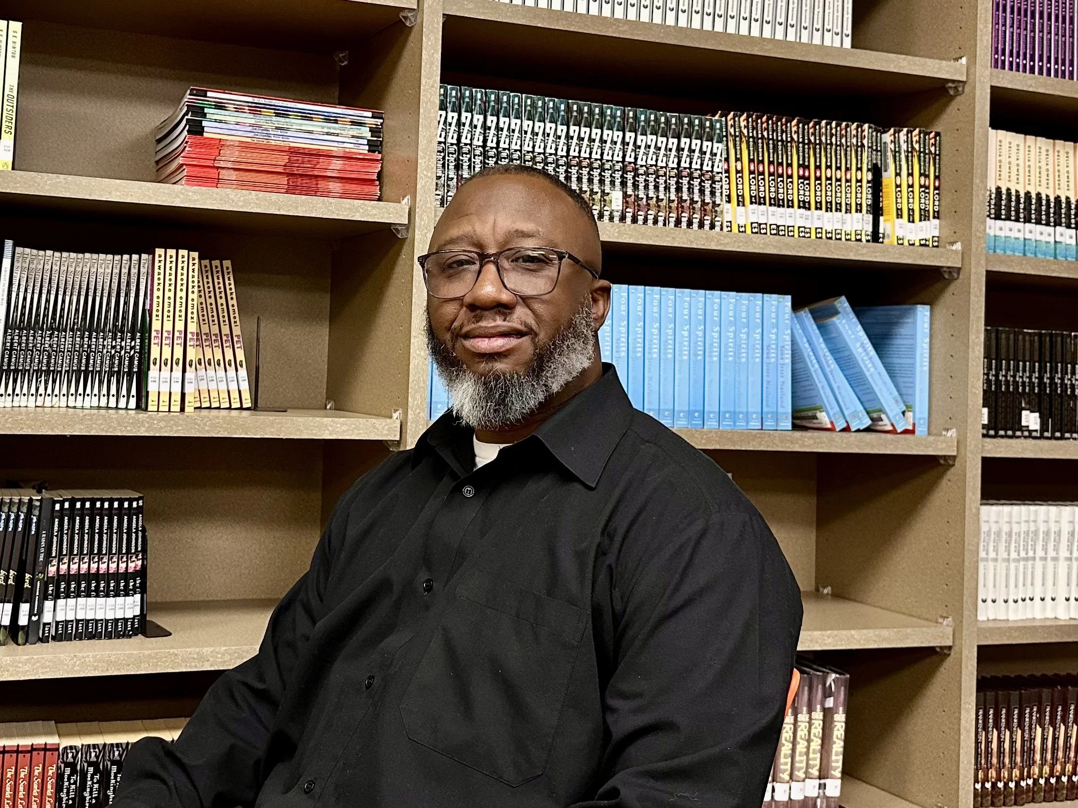 A man with glasses, a black shirt, and a gray beard sitting in front of bookshelves filled with graphic novels and comic books.