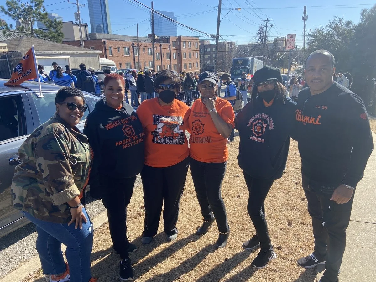 Group of six people standing outdoors in a sunny urban area with a crowd in the background. They are dressed in clothing supporting Douglass High School and alumni, with some wearing matching black and orange shirts and hoodies. A car with a Trojan f