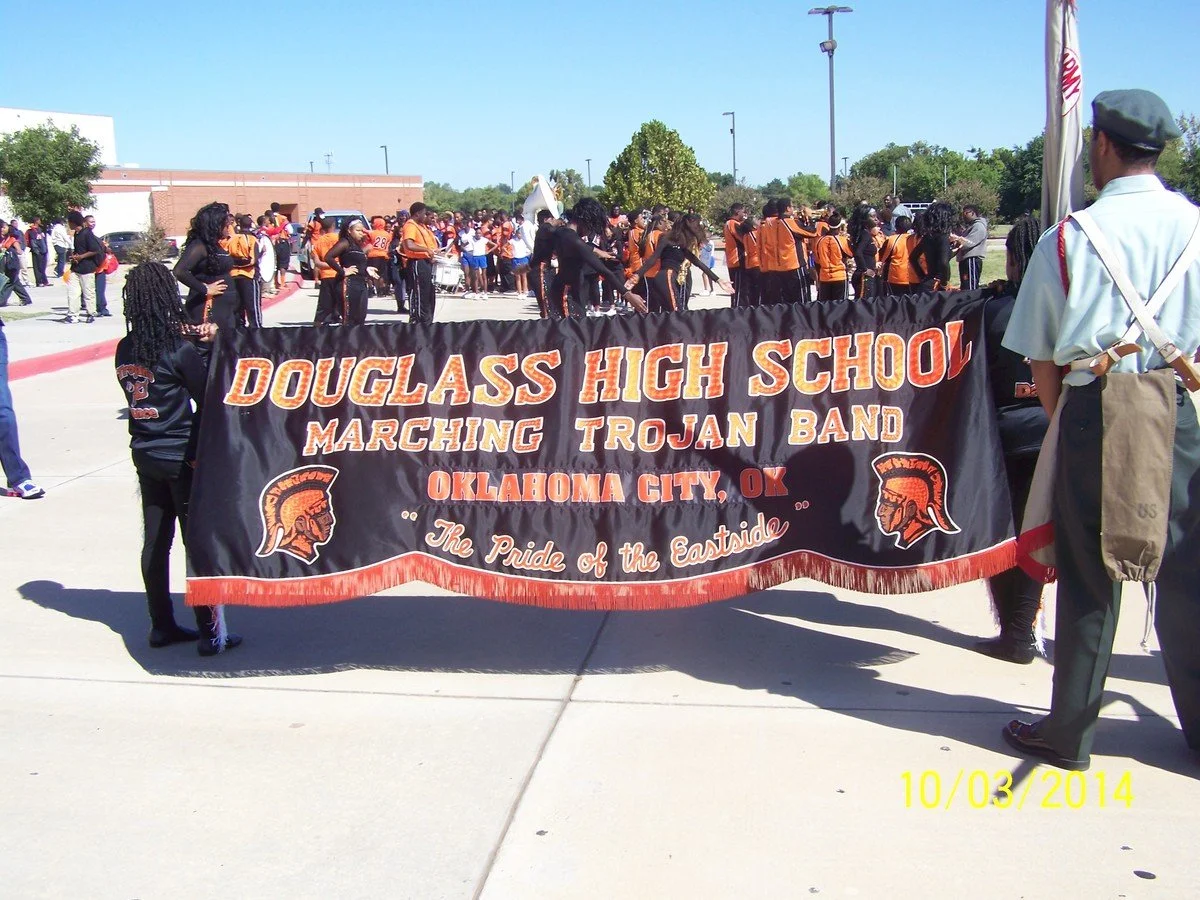 A parade with students from Douglass High School marching band in Oklahoma City; they are holding a large banner and many students are dressed in black and orange uniforms.