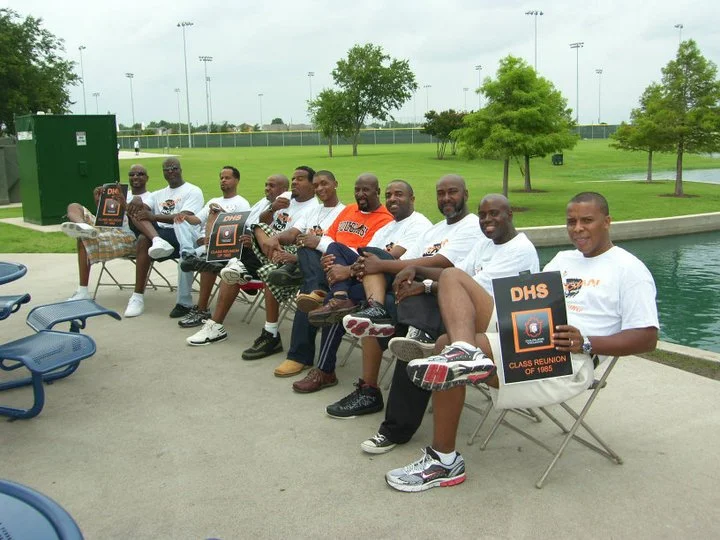 A group of nine men sitting on chairs by a pond, holding reunion flyers, with a park, trees, and sports field in the background.