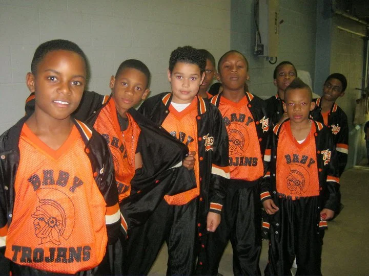 Group of seven young girls in orange and black basketball jerseys and jackets, standing in a sports facility.