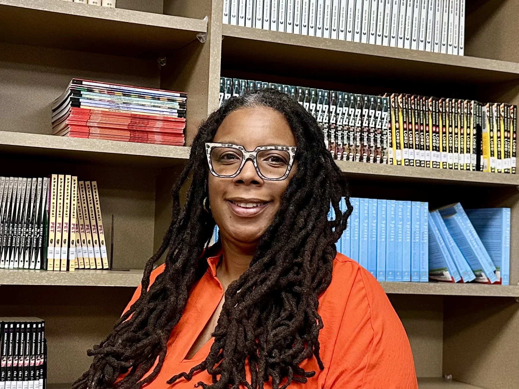 A woman with long dreadlocks, wearing glasses with a leopard print pattern and an orange shirt, smiling in front of a bookshelf filled with books and magazines.