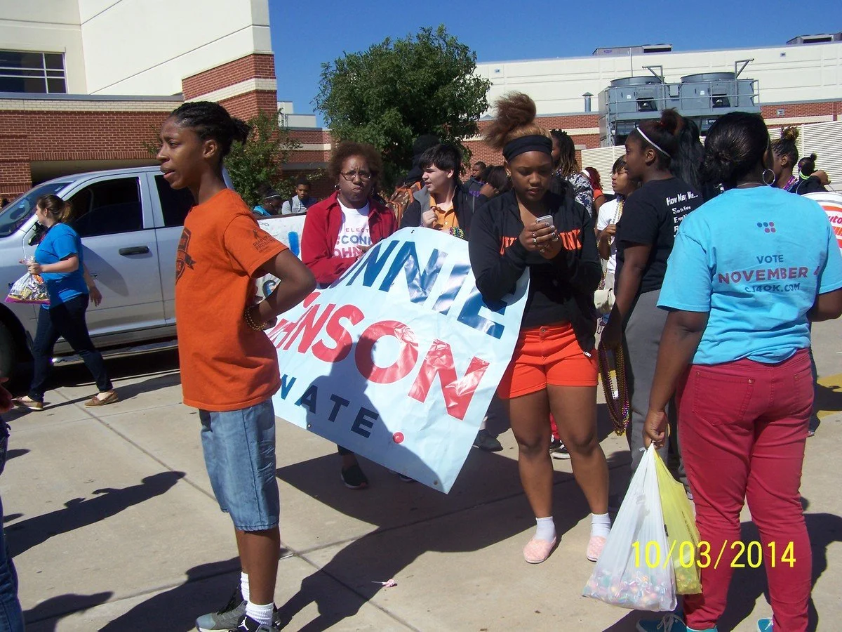 Group of people protesting on the sidewalk, holding a large sign that reads 'End Violence Against Women' and wearing shirts promoting voting in November 2014.