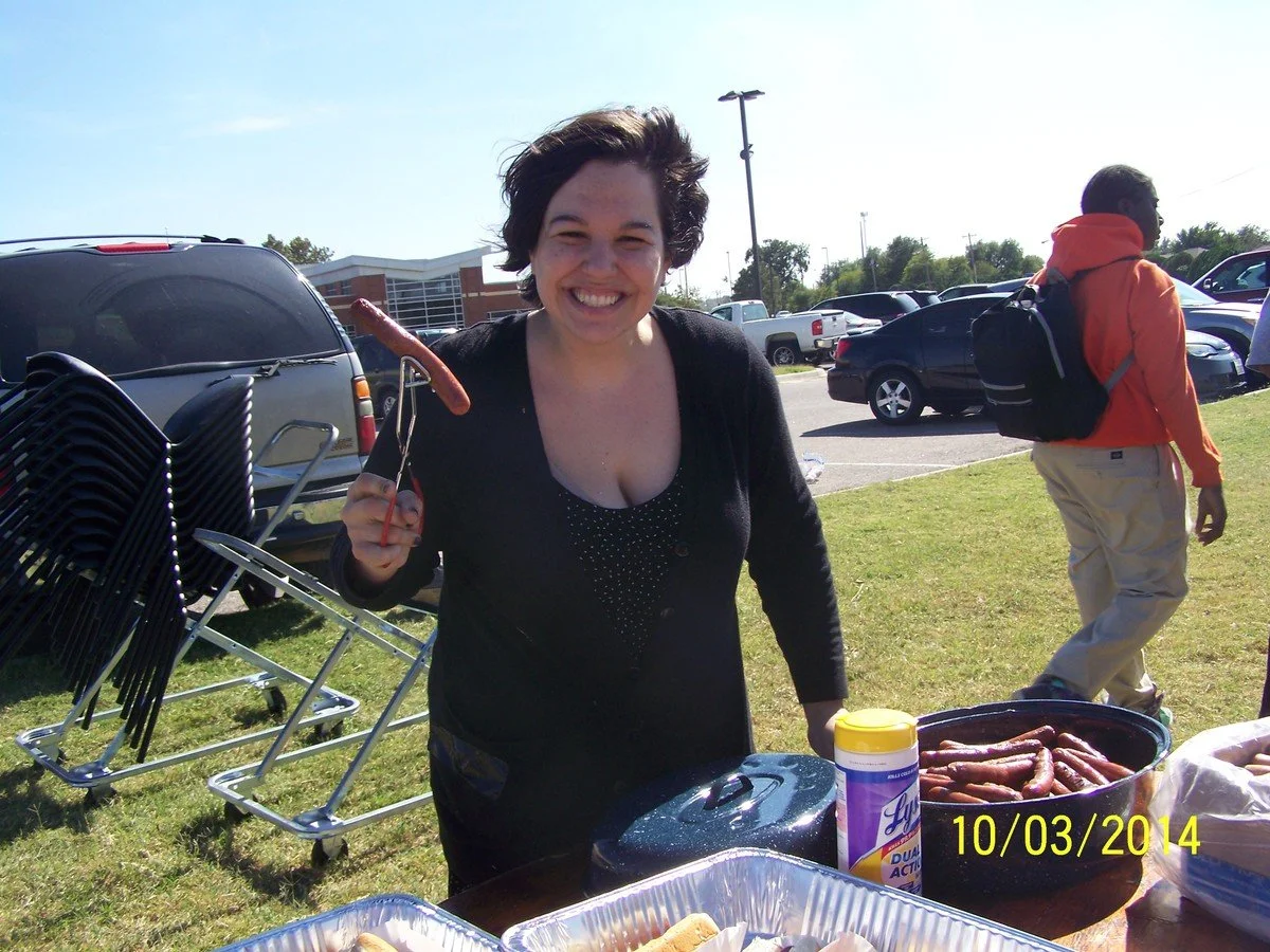 A woman smiling and holding a hot dog in a park at a barbecue event. There are tables, chairs, and parked cars in the background on a sunny day.