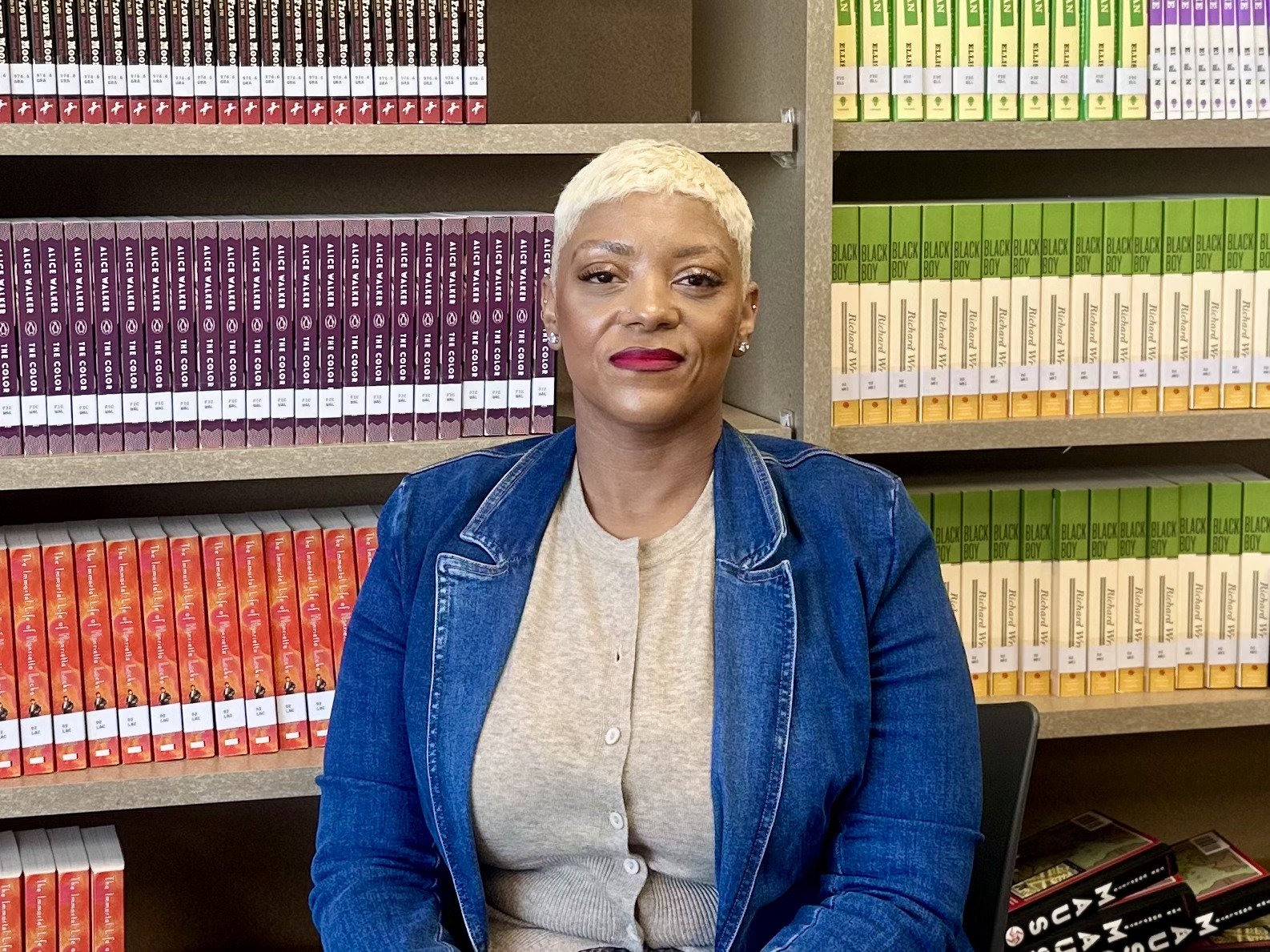 A woman with short platinum blonde hair, wearing a blue denim jacket and light gray top, sitting in front of bookshelves filled with colorful books.