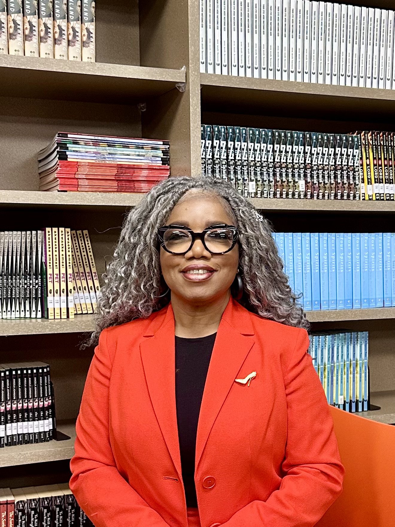 A woman with gray curly hair and glasses smiling, wearing a red blazer with a bird brooch, sitting in front of bookshelves filled with colorful books.