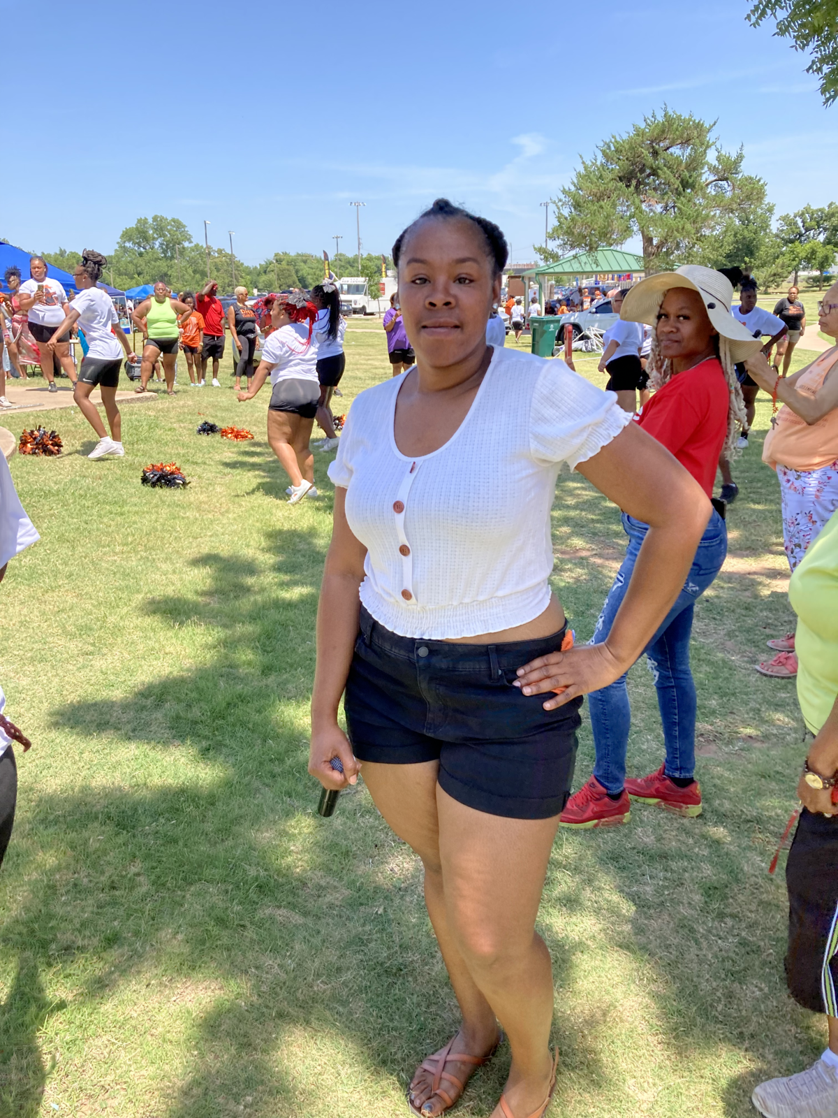 A woman standing outdoors on a grassy field during a sunny day, with many people participating in a group activity or event in the background, some stretching or dancing.