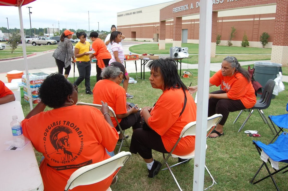 Group of women wearing matching orange shirts sitting and talking under a canopy outside a school building, with children lined up nearby and a table with speakers and refreshments.
