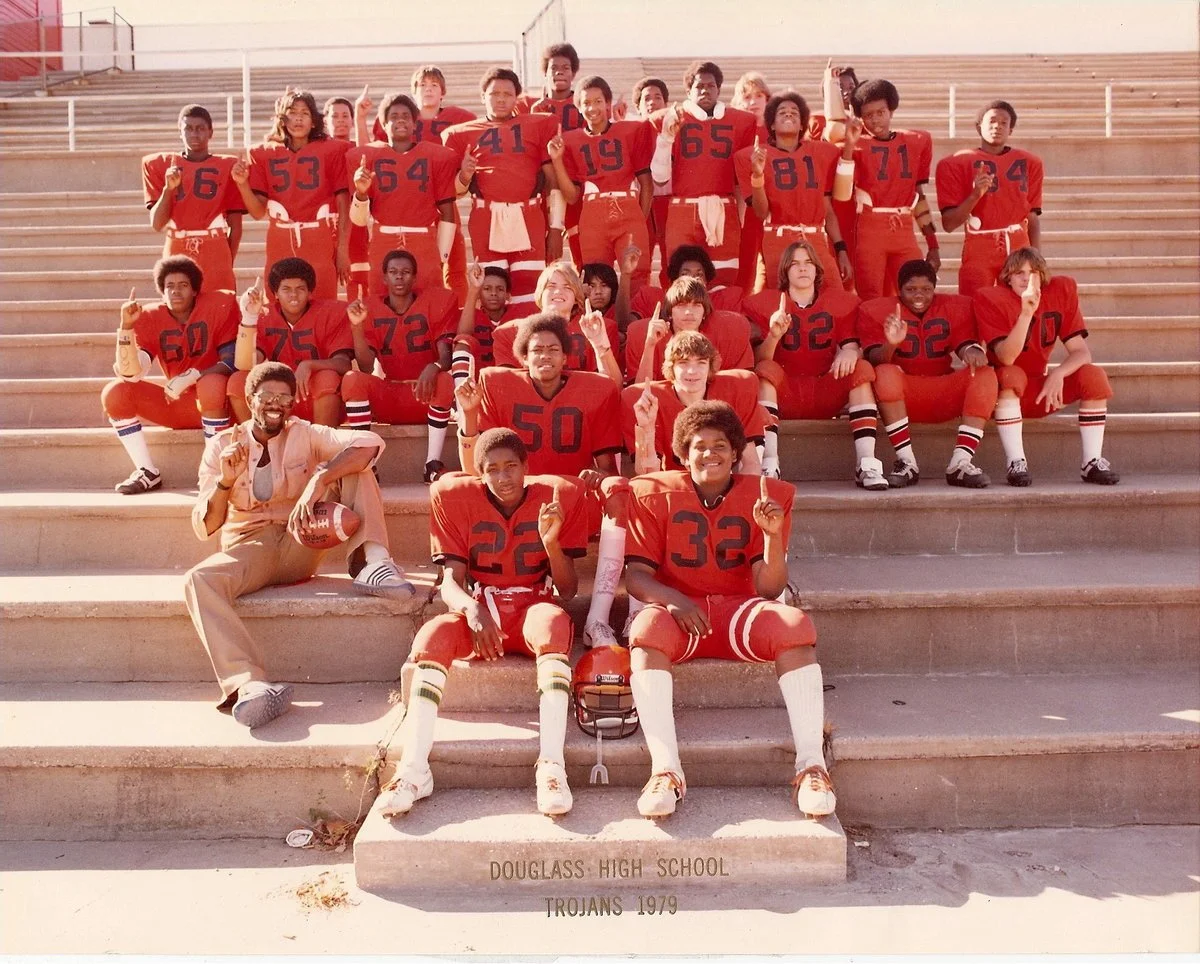 Group of high school football players in red uniforms on bleachers, with coach, in 1979 at Douglass High School Trojans.