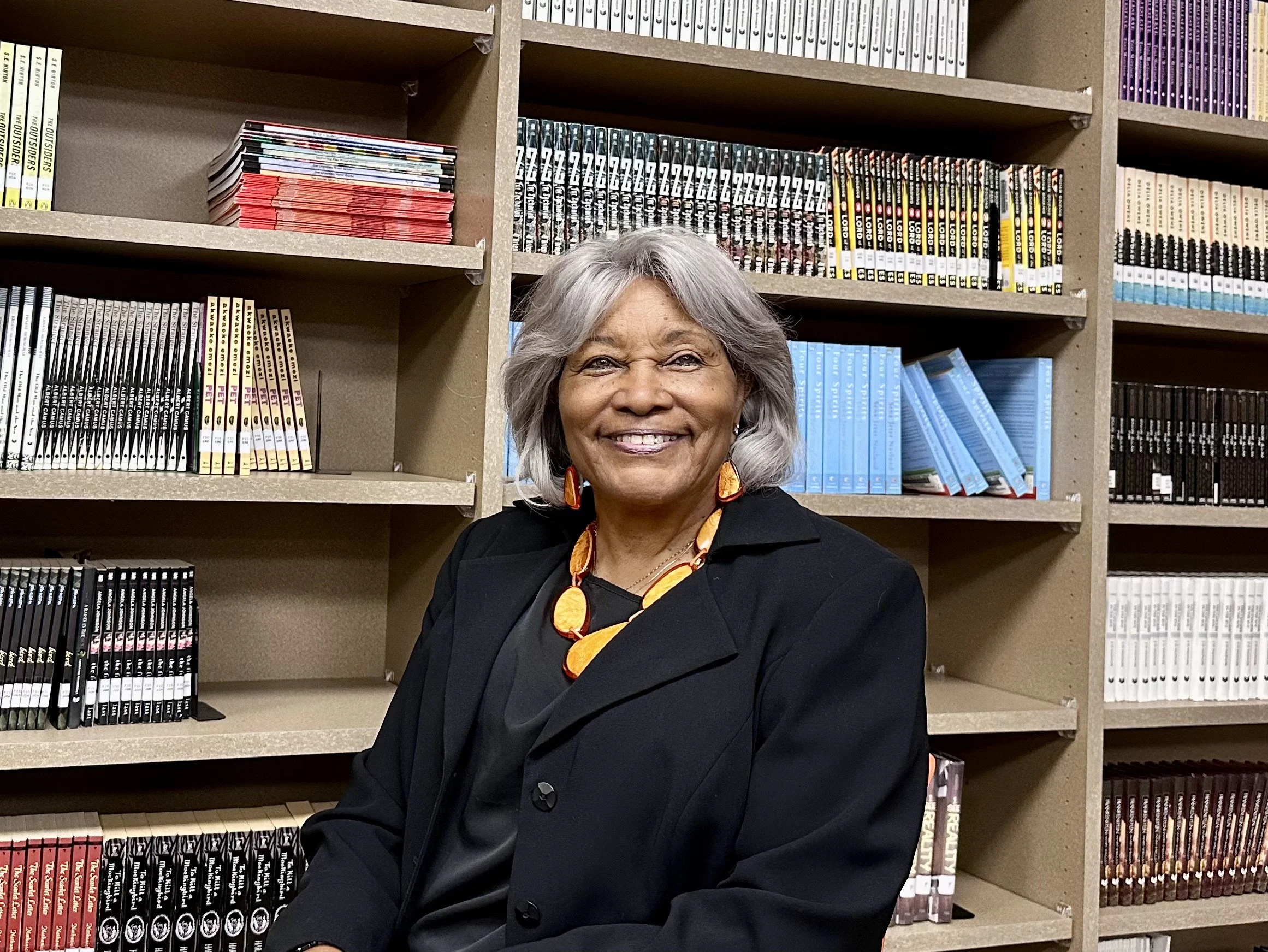 An elderly woman with gray hair, wearing a black blazer, colorful necklace, and matching earrings, smiling while sitting in front of a bookshelf filled with various books and magazines.