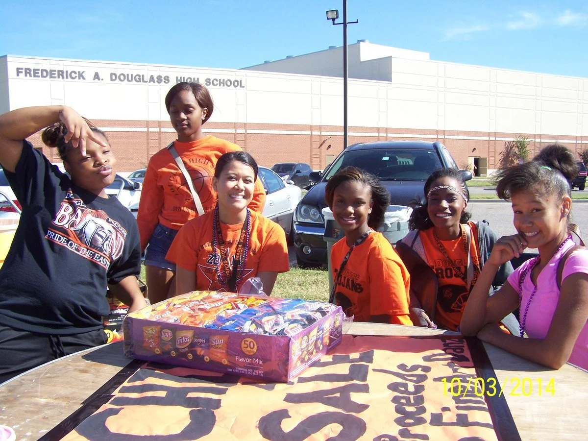 A group of six young girls gathered around a table outside of Frederick A. Douglass High School, with a large box of snacks on the table, smiling and posing for the camera on a sunny day.