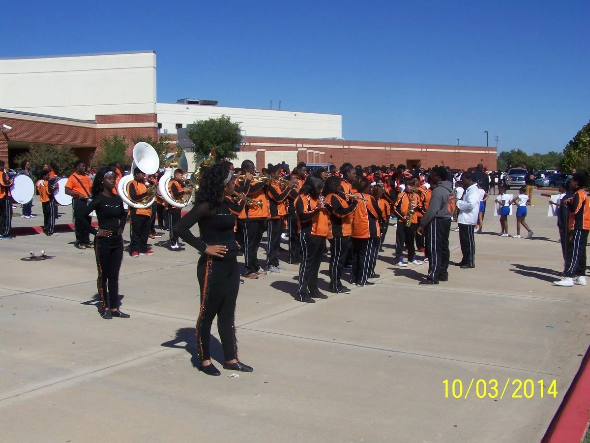 A school marching band performing outdoors in a parking lot, with students dressed in black and orange uniforms, and cheerleaders in blue and white uniforms in the background.