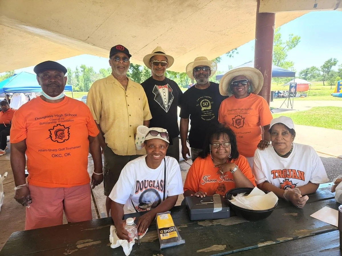 Group of eight people, mostly women, gathered outdoors under a pavilion, some wearing orange T-shirts with event information about a high school golf tournament and scholarship foundation, with a park and greenery in the background.