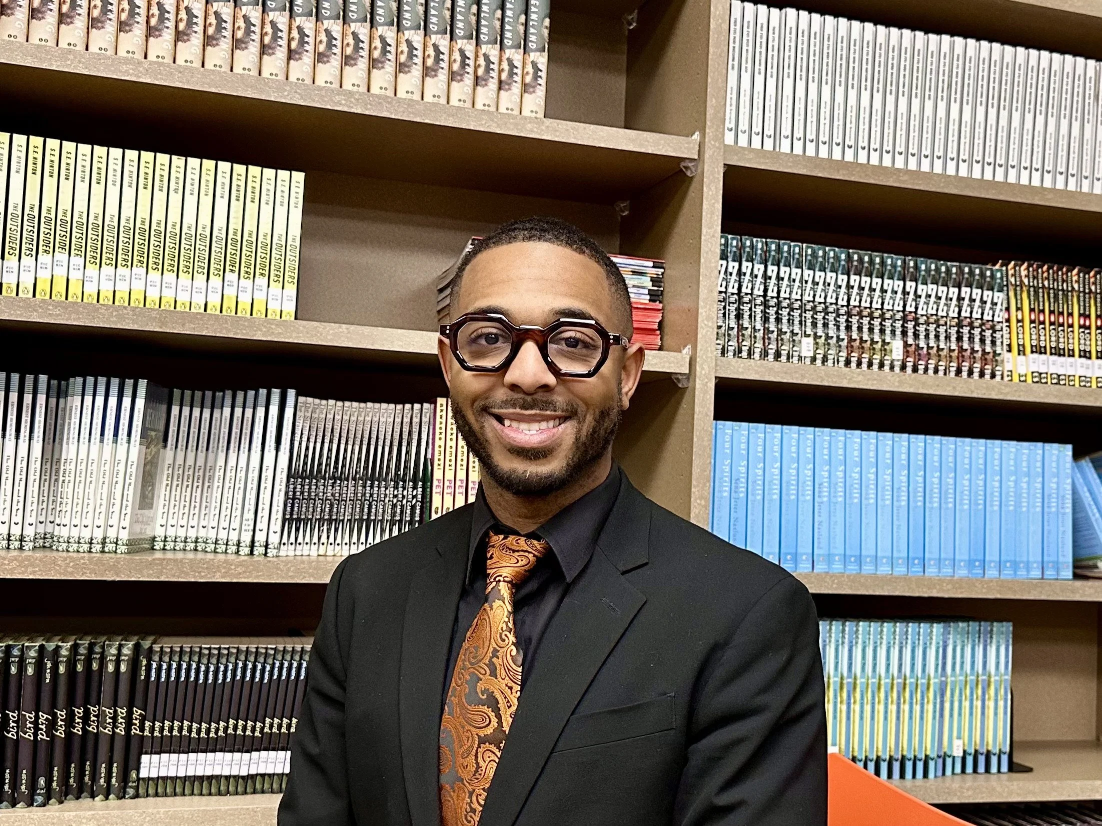 A smiling man wearing a black suit, paisley orange tie, and glasses standing in front of bookshelves filled with comic books.