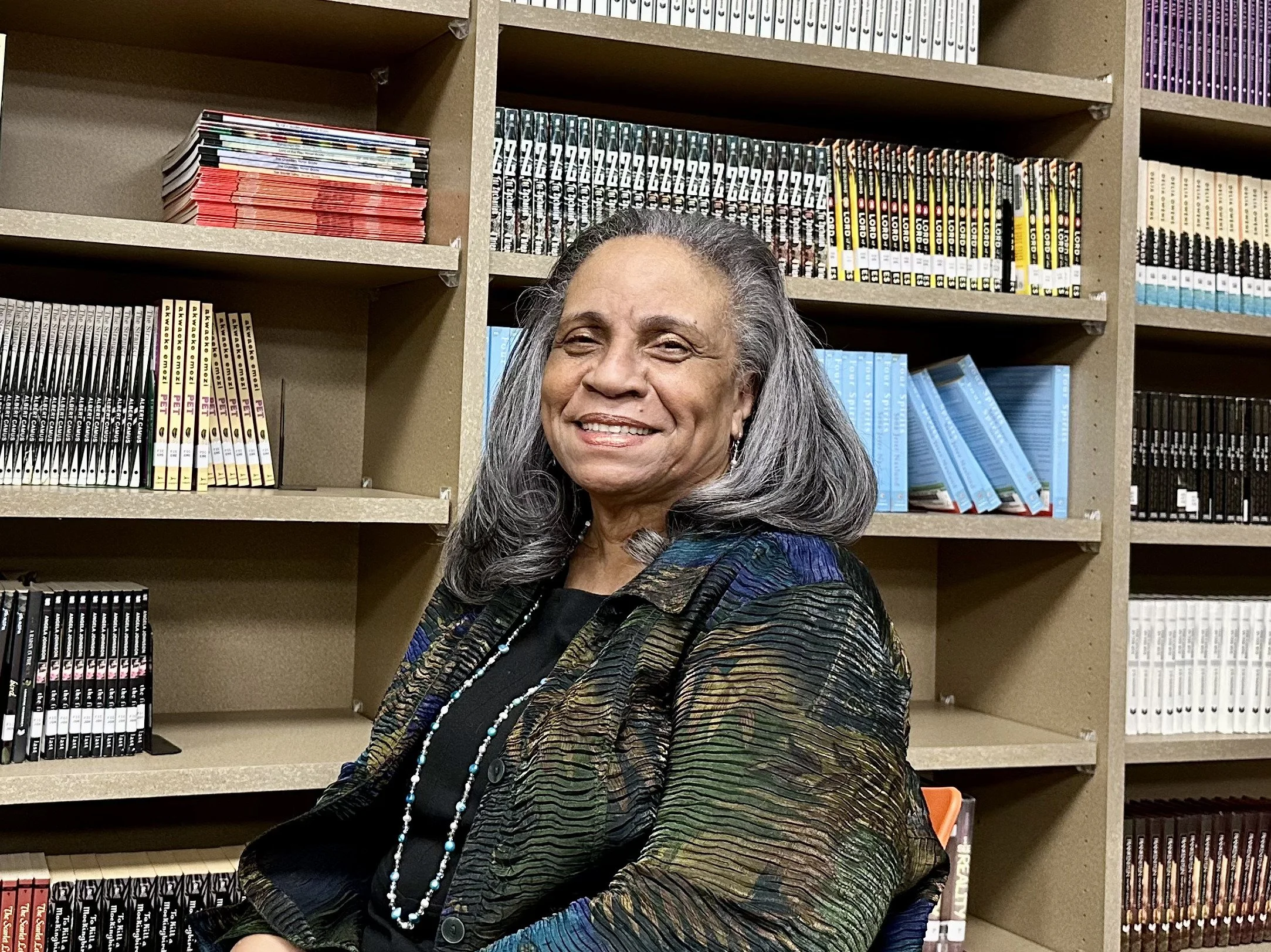 A smiling woman with gray hair seated in front of a bookshelf filled with colorful books.