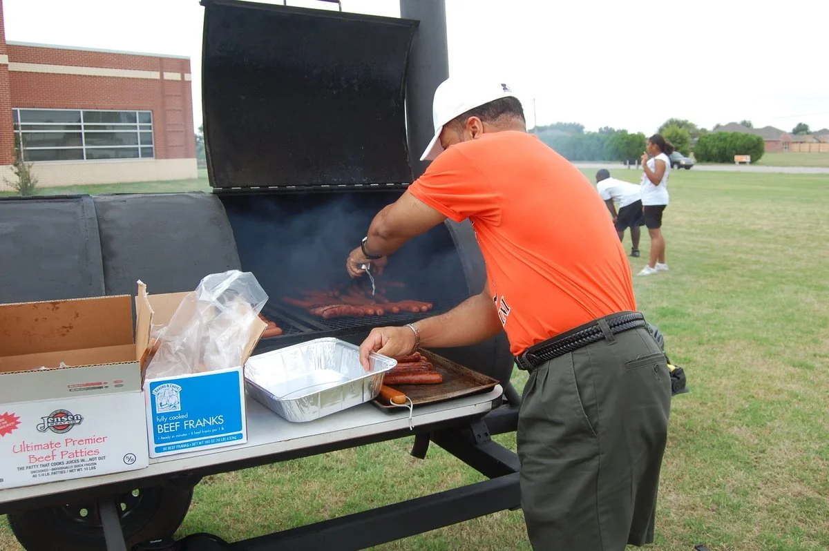 A man in a white cap and orange shirt grills hot dogs on a barbecue grill outdoors, with a few people in the background and a grassy field.