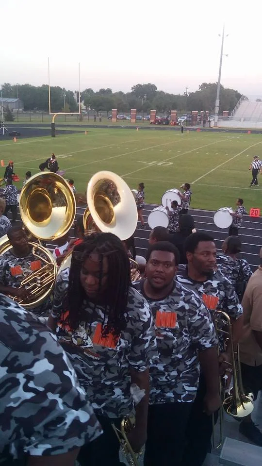 Band members in camouflage shirts holding musical instruments at a football field, with cheerleaders in the background.