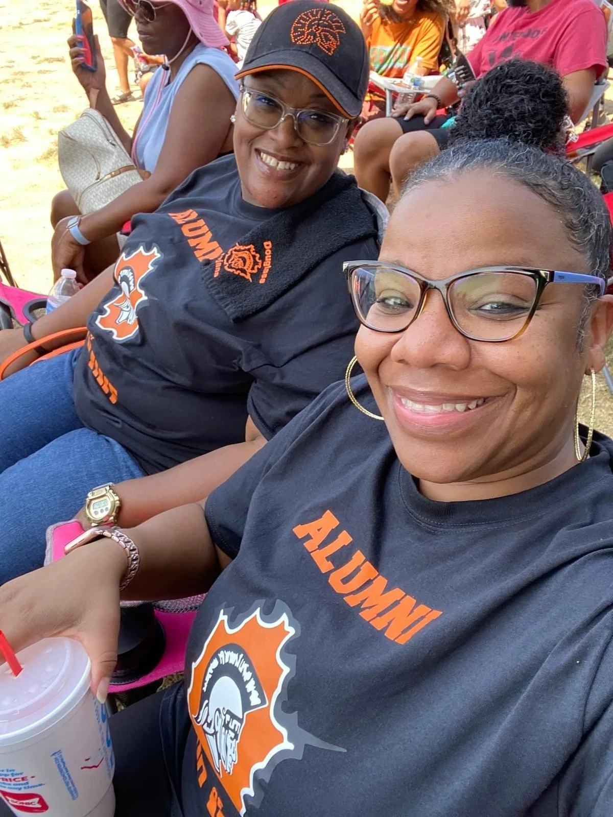 Two women smiling at a crowded outdoor event, wearing black T-shirts with orange text and a logo. One holds a drink with a straw, and others are seated behind them.