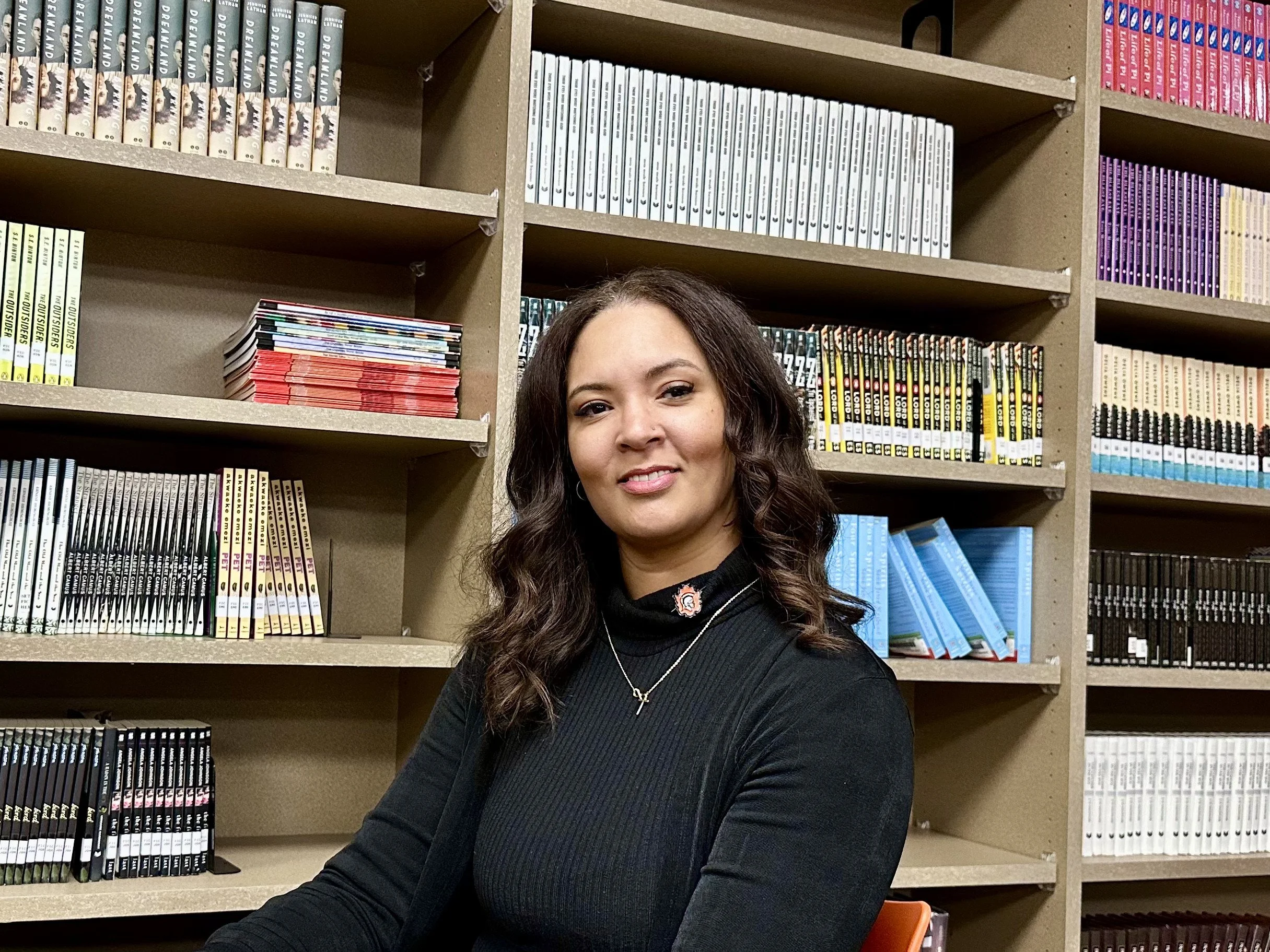 A woman with dark, wavy hair wearing a black top and a necklace, sitting in front of bookshelves filled with textbooks and magazines.