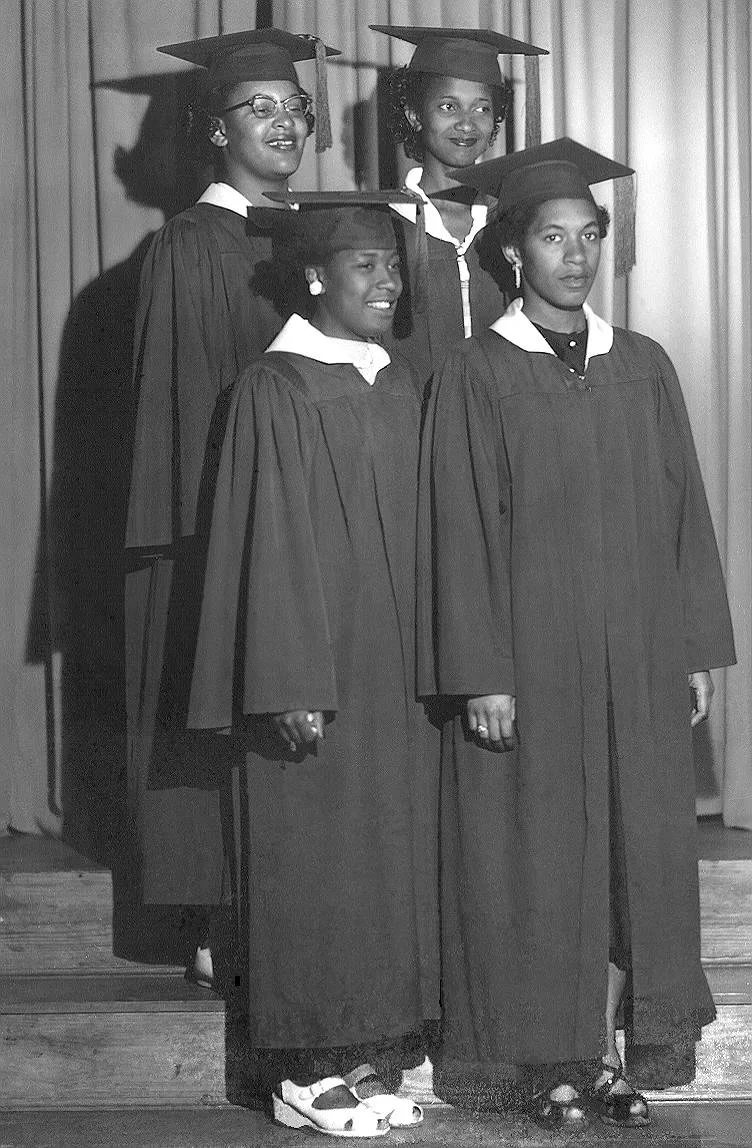 Black and white photograph of four women in graduation gowns and caps standing on a stage in front of curtains. They are smiling and looking at the camera.