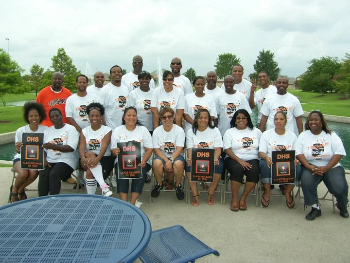 Group of adults outdoors by a water fountain, some holding signs that read 'DHS Class Reunion 2000', posing for a photo on a cloudy day.
