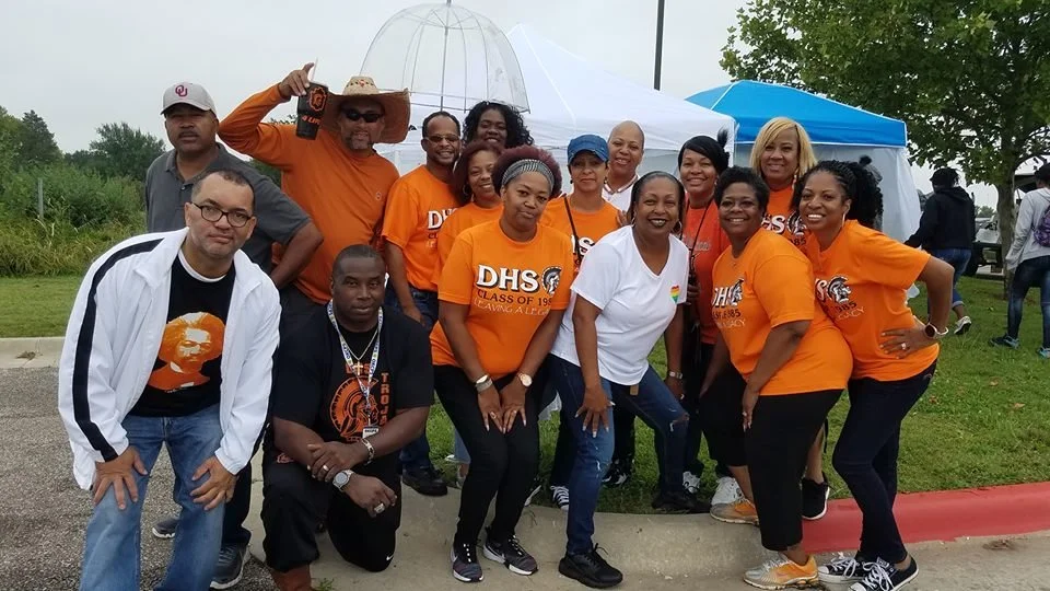 Group of people, mostly women, posing outdoors at an event, many wearing orange T-shirts with 'DHS Class of 1984' printed on them, some in casual attire, with tents and trees in the background.