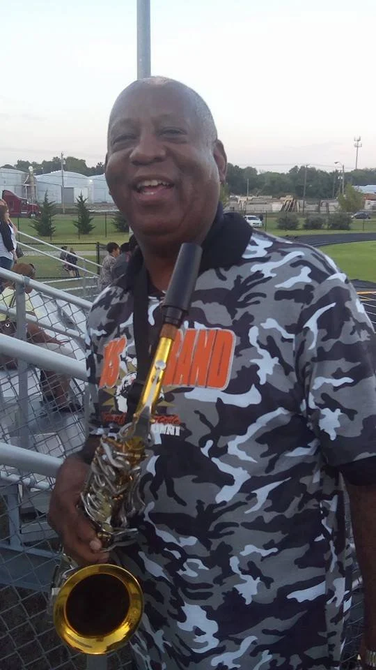 A smiling man holding a saxophone at an outdoor event with bleachers and people in the background.