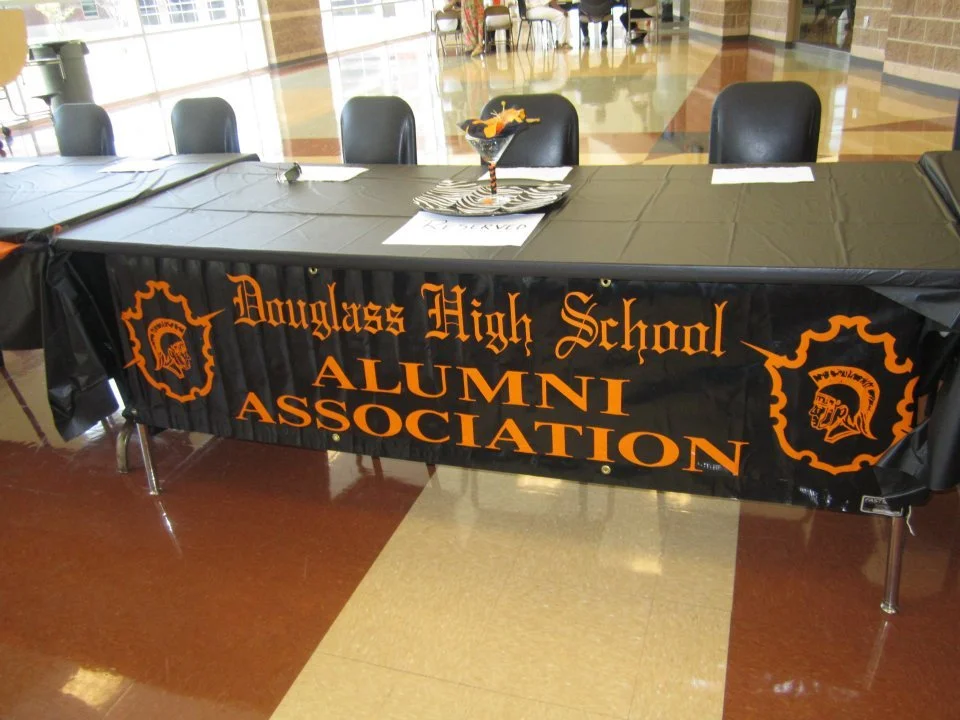 Table with a black tablecloth displaying 'Doughlas High School ALUMNI ASSOCIATION' in orange text, and black chairs around it. A centerpiece with orange and black decorations is on the table, in a large indoor space with tiled floors and large window