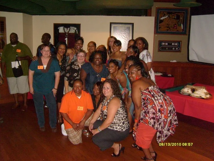 Group of women and a few men posing together in a restaurant or event space, some wearing name tags, with artwork on the walls and a long table with a cake in the background.